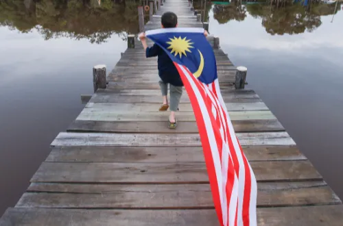 Child walks down a wooden pier holding a Malaysian flag, surrounded by calm water and greenery. The mood is peaceful and patriotic.